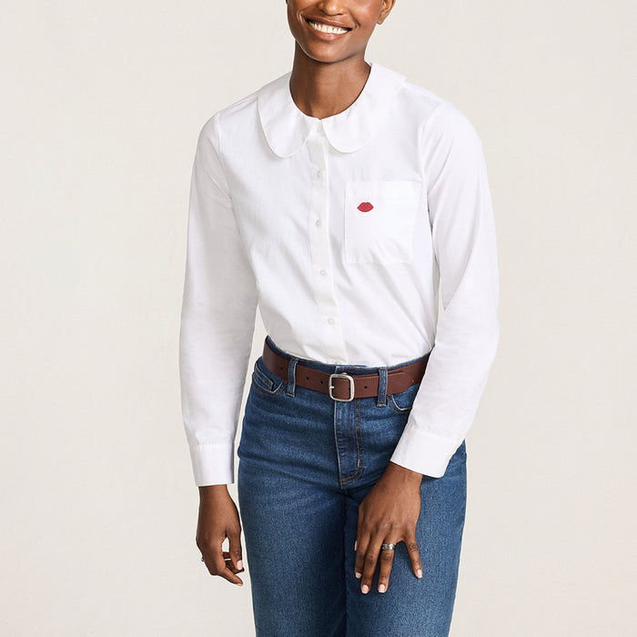 Women wearing a white cotton shirt with a round peter pan collar and red embroidered lip on the pocket tucked into denim jeans against a beige background