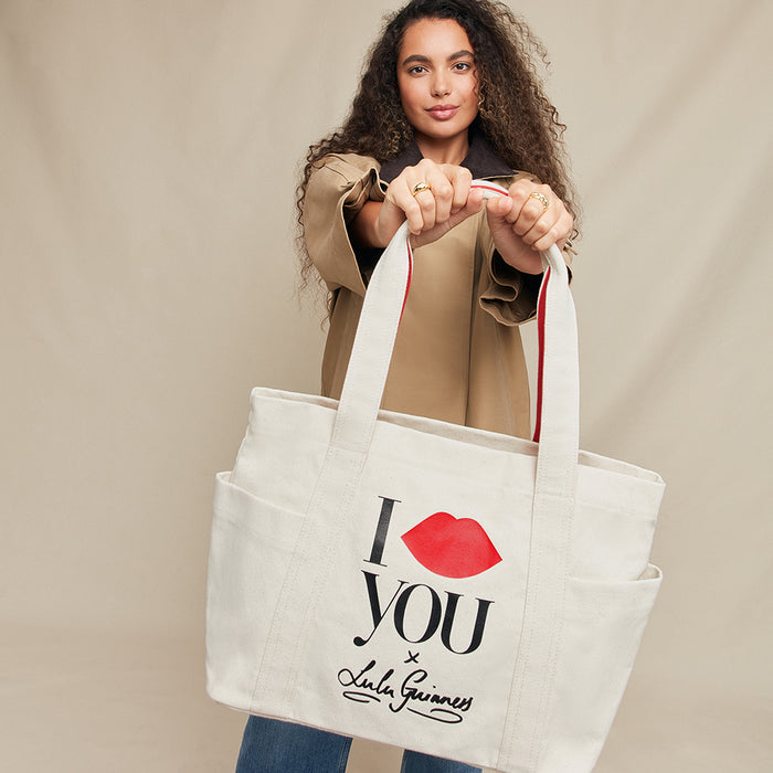 Woman holding a tote bag with 'I lip you x Lulu Guinness' text and red lips design on a beige background